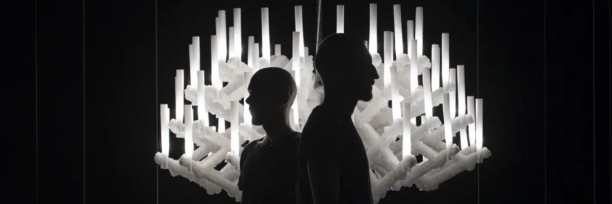 Zwei Schattengestalten in einem dunklen Raum vor einem hellen Kronleuchter