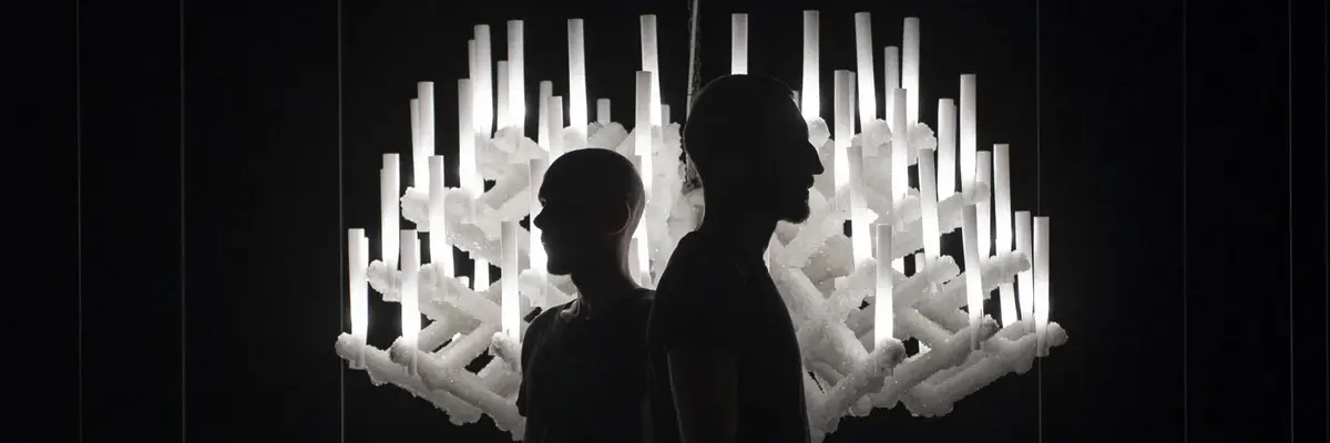 Zwei Schattengestalten in einem dunklen Raum vor einem hellen Kronleuchter