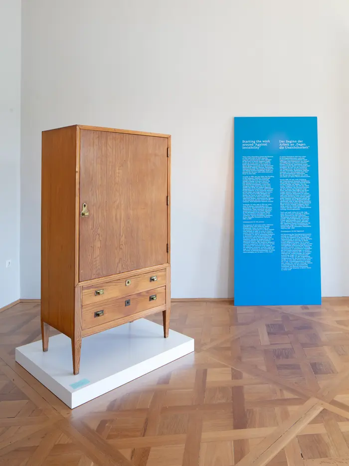 Ausstellungsansicht mit einem hölzernem Schrank und einer Informationstafel