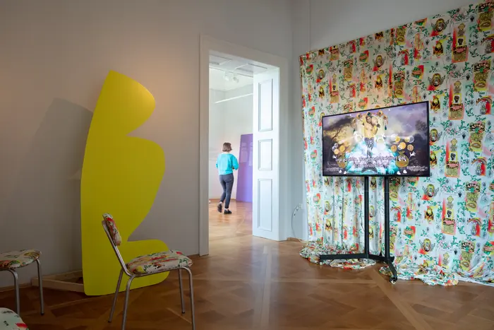Ausstellungsansicht eines Raumes mit einem Video vor einem bunten Wandbehang mit Blick in den nächsten Raum