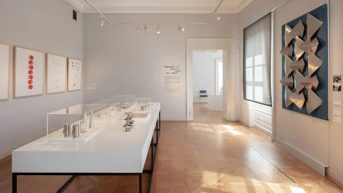 Ausstellungsansicht eines Präsentationstisches auf dem Silberwaren in Vitrinen ausgestellt sind und Bildern 