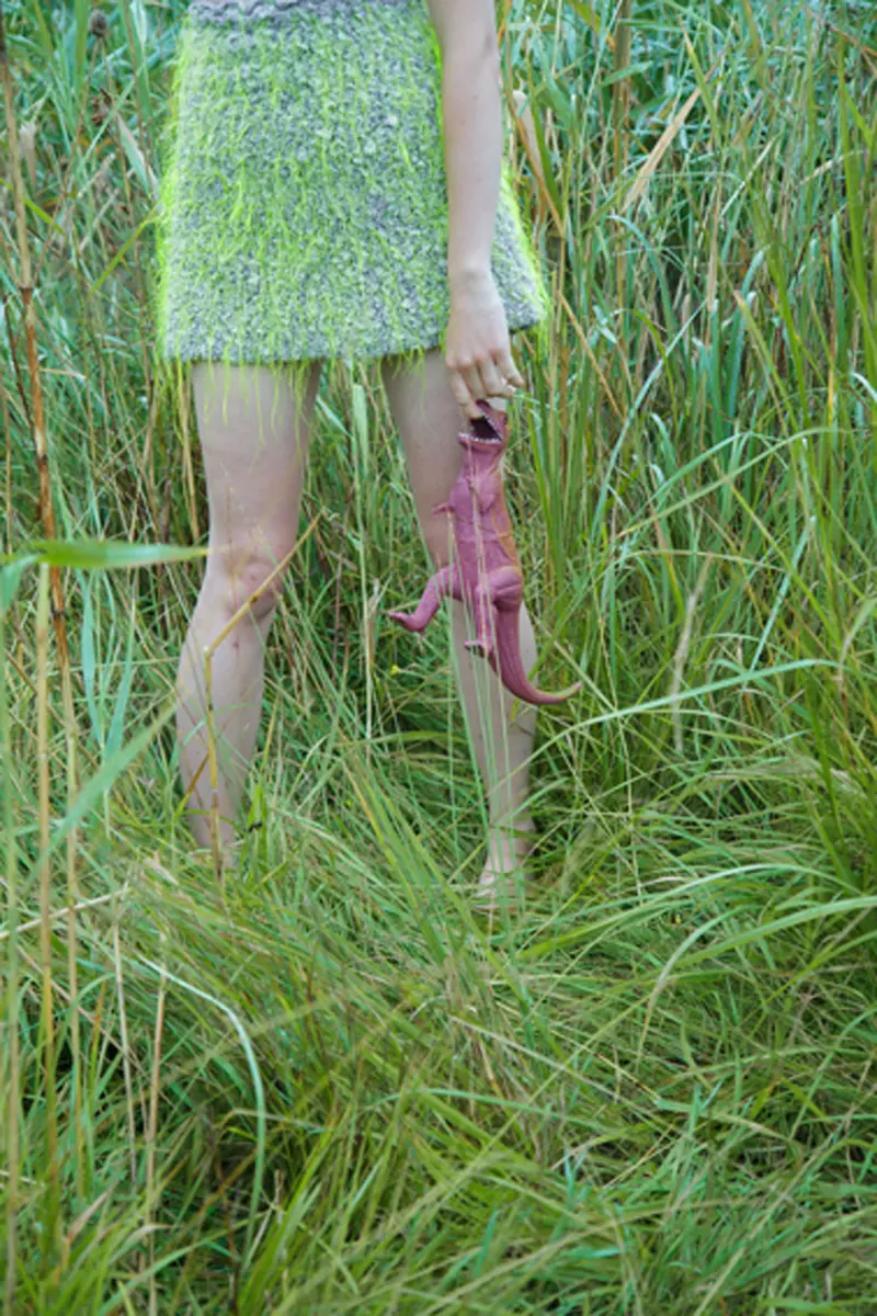 Unterkörper einer Person in Kleid, steht im Gras, lässt Dinosaurierfigur am Finger hängen