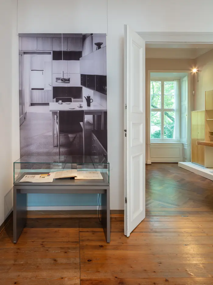 Ausstellungsansicht mit einer Vitrine gefüllt mit zwei Büchern und einer Fotografie einer Inneneinrichtung