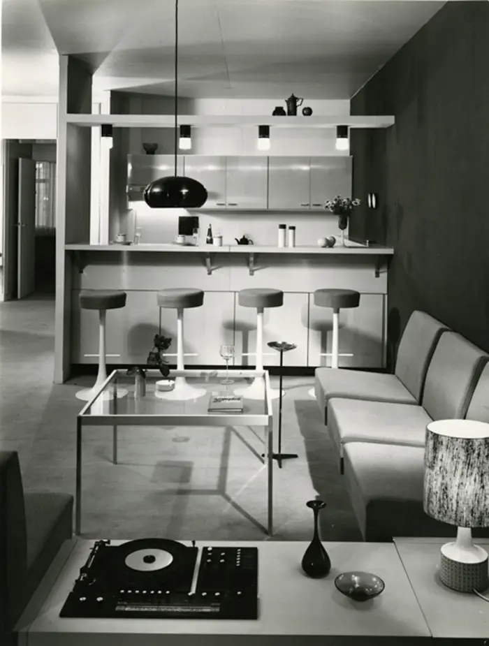 Schwarz-Weiß-Foto eines Wohnzimmers mit Durchbruch zur Küche im Stil der 1960er/ 1970er Jahre