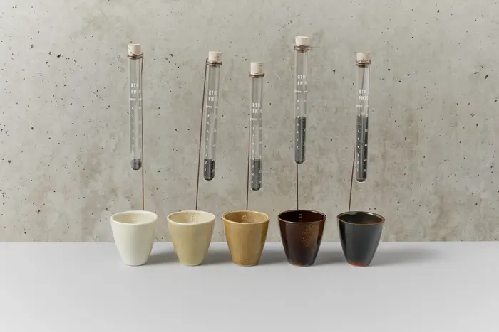 Fünf kleine Becher in verschiedenen Braunstufen, über denen jeweils ein Reagenzglas mit brauner Flüssigkeit angebracht ist